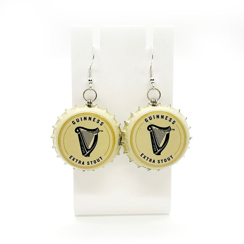 May include: A pair of earrings made from repurposed Guinness beer bottle caps. The caps are gold with black lettering that reads "GUINNESS EXTRA STOUT" and a black harp logo. The earrings are attached to silver hooks.