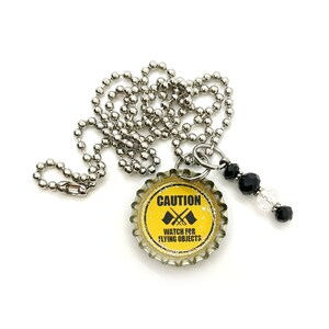 May include: A silver chain necklace with a yellow bottle cap pendant that says "CAUTION WATCH FOR FLYING OBJECTS" with a black and white graphic of crossed flags. The pendant is attached to a silver ring with black and clear crystal beads.
