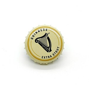 May include: A Guinness Extra Stout bottle cap. The cap is cream-colored with a gold rim and text. The center features a black harp logo and the words "GUINNESS" and "EXTRA STOUT". The cap is metal and has a crimped edge.