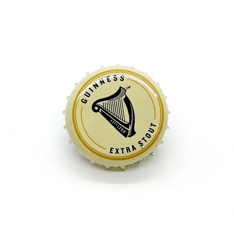 May include: A Guinness Extra Stout bottle cap. The cap is cream-colored with a gold rim and text. The center features a black harp logo and the words "GUINNESS" and "EXTRA STOUT". The cap is metal and has a crimped edge.