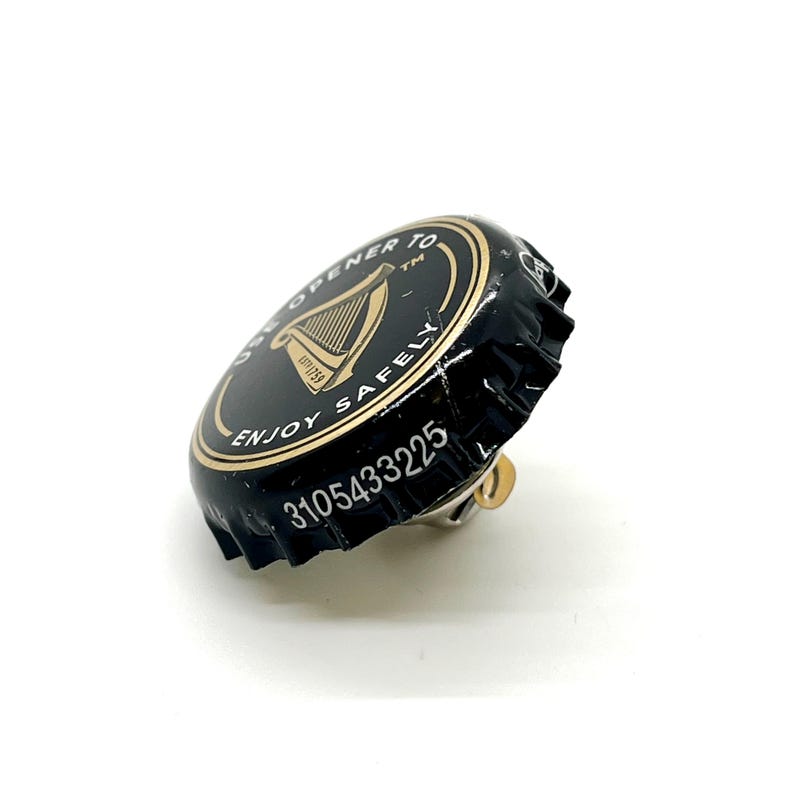 May include: A black and gold Guinness bottle cap with the text "ONE OPENER TO ENJOY SAFELY" and a harp logo. The cap has a silver pin attached to the back. The serial number 3105433225 is printed on the side.