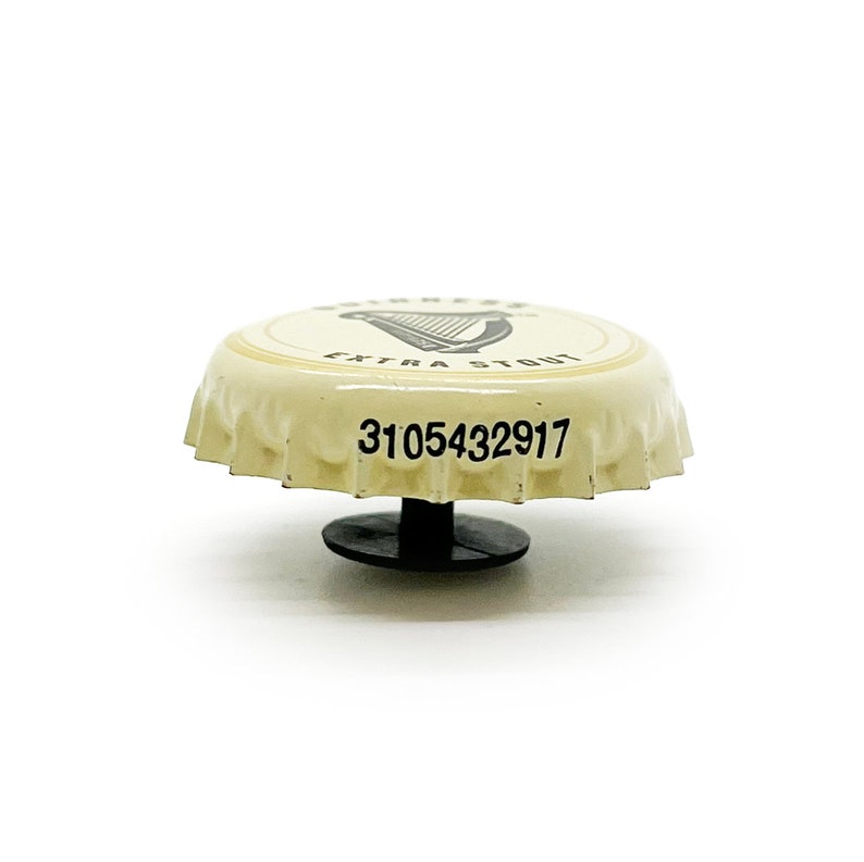 May include: A white bottle cap with a black harp logo and the text "Guinness Extra Stout". The bottle cap has a black plastic ring on the bottom and the number "3105432917" printed on the top.