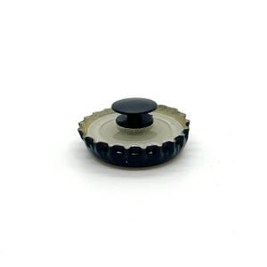May include: A black bottle cap with a black knob on top. The bottle cap has a silver interior and a dark blue exterior. The knob is round and appears to be made of plastic. The bottle cap is sitting on a white surface.