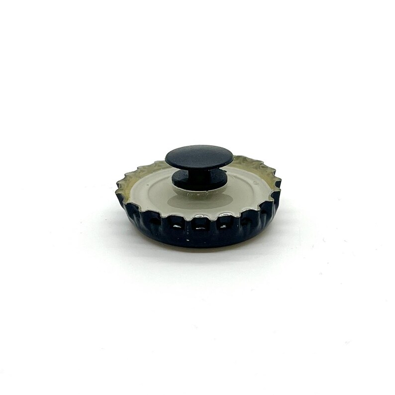 May include: A black bottle cap with a black knob on top. The bottle cap has a silver interior and a dark blue exterior. The knob is round and appears to be made of plastic. The bottle cap is sitting on a white surface.