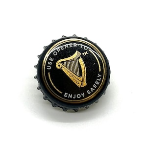 May include: A black bottle cap with a gold harp design and the text "USE OPENER TO ENJOY SAFELY". The cap has a gold rim and is from the Guinness brand. The cap is on a white background.