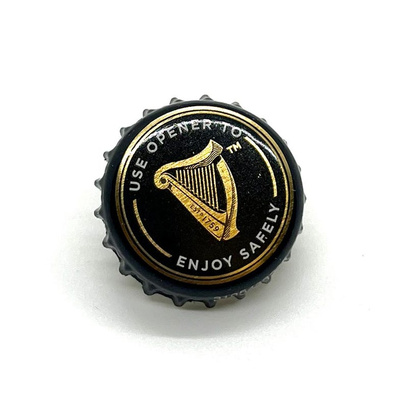 May include: A black bottle cap with a gold harp design and the text "USE OPENER TO ENJOY SAFELY". The cap has a gold rim and is from the Guinness brand. The cap is on a white background.