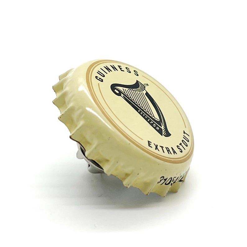 May include: A Guinness Extra Stout bottle cap. The cap is off-white with a black harp logo and text. The text includes "GUINNESS", "ESTD 1759", and "EXTRA STOUT". The cap has a metallic silver edge.