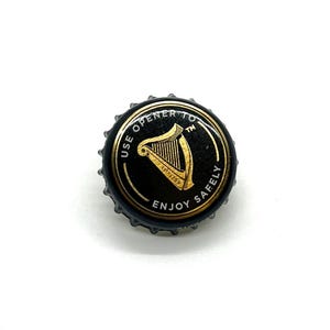 May include: A black bottle cap with gold lettering and a gold harp design. The text on the cap reads "USE OPENER TO ENJOY SAFELY". The harp design includes the text "EST 1759".