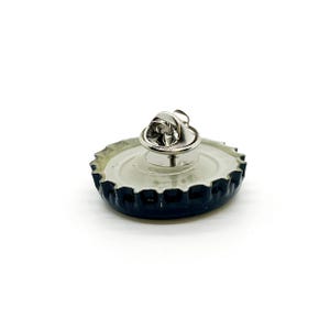 May include: A bottle cap lapel pin with a silver pin back. The bottle cap is black with a silver rim and a white interior. The pin is a unique accessory.