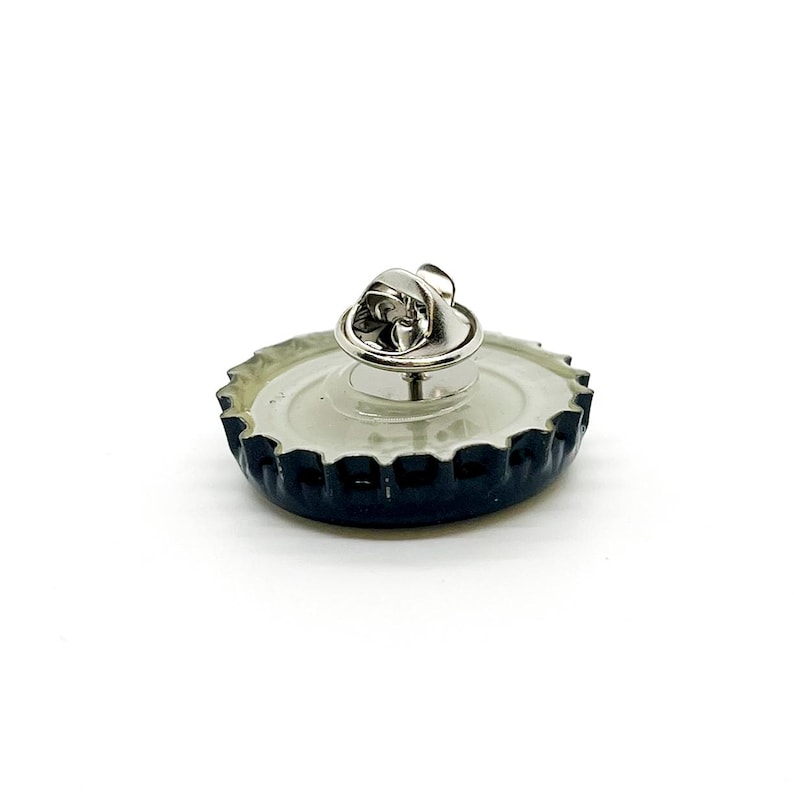May include: A bottle cap lapel pin with a silver pin back. The bottle cap is black with a silver rim and a white interior. The pin is a unique accessory.