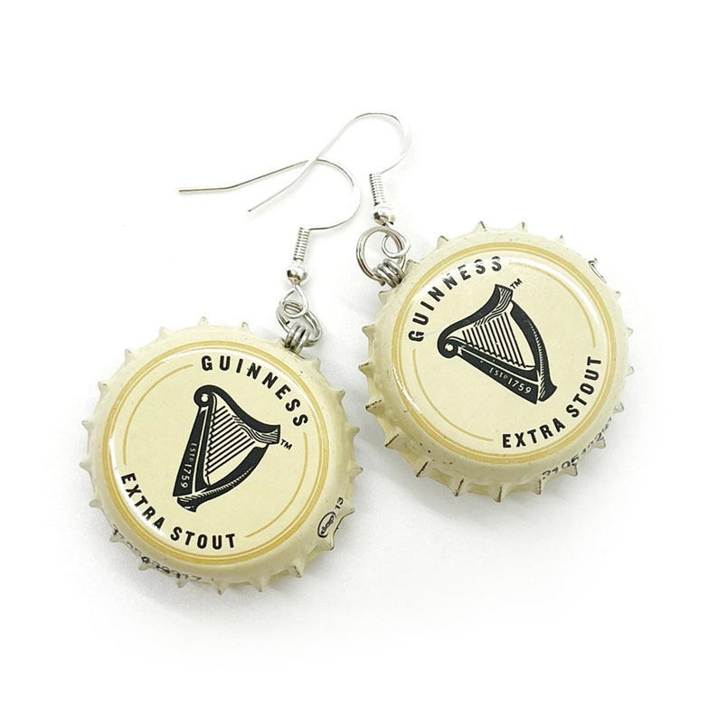 May include: A pair of earrings made from repurposed Guinness bottle caps. The caps are cream colored with black lettering and a harp logo. The earrings are silver and have a hook closure.