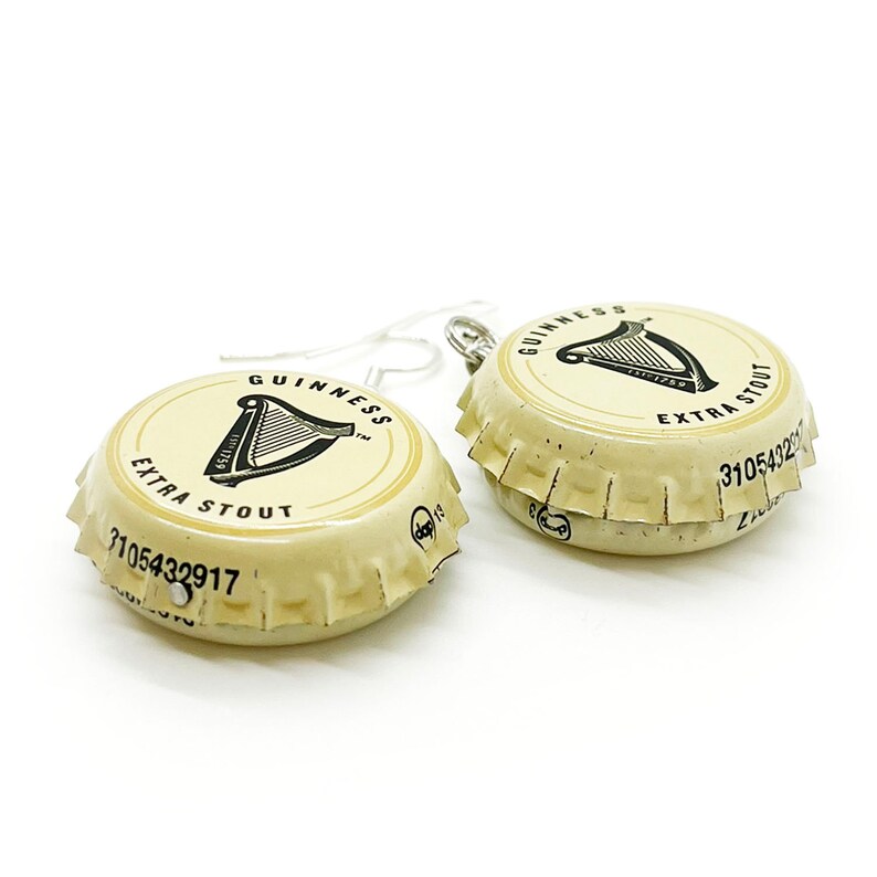 May include: A pair of earrings made from Guinness beer bottle caps. The caps are cream colored with black lettering that reads "GUINNESS EXTRA STOUT" and feature a black harp logo. The earrings are attached to silver hooks.