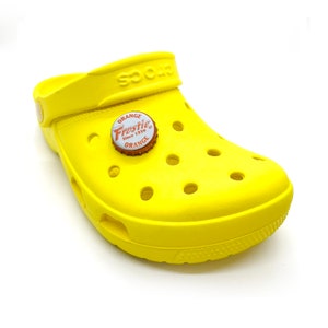 May include: Yellow clog with a round, orange and white Frostie bottle cap charm. The charm has the text "Orange Frostie Since 1939 Orange" on it.