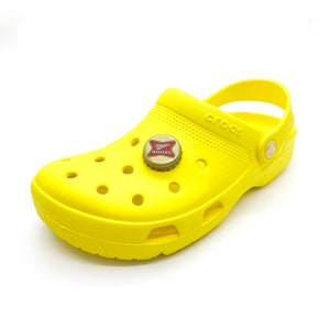 May include: Bright yellow Crocs shoe with a bottle cap charm. The bottle cap has a red and white design with the words "Miller High Life". The shoe has multiple ventilation holes and the word "Crocs" is visible on the side.