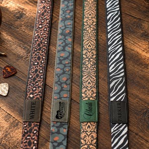 Può includere: Quattro tracolle per chitarra con motivi distinti: una stampa leopardata marrone con un'etichetta che riporta la scritta WHITE, una stampa leopardata grigia e arancione con un'etichetta che riporta Griffin, una stampa damascata verde con un'etichetta che riporta Clark e una stampa zebrata bianca e nera con un'etichetta che riporta PARKER.