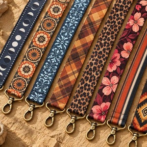 May include: Eight long, flat fabric bag straps arranged side-by-side, each featuring a unique pattern including moon phases, floral motifs, plaid, leopard print, and geometric designs, all finished with antique brass-toned metal swivel hooks.
