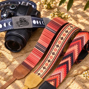 Op de afbeelding: Een zwarte spiegelreflexcamera met een donkerblauwe camerariem met een wit maanfasenpatroon en een grijze patch met de tekst Collins. Daarnaast liggen twee extra camerariemen met rode, oranje en zwarte geometrische en gestreepte patronen.