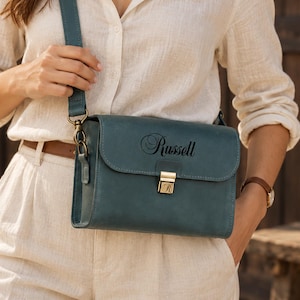 Peut inclure: Un sac bandoulière en cuir bleu sarcelle doté d'un fermoir en métal doré avec une lettre R gravée. Le rabat avant est personnalisé avec le nom Russell en police cursive noire. Le sac comprend une bandoulière réglable assortie.