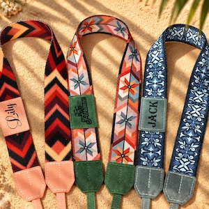 May include: Three camera straps with distinct patterns. The first features red, orange, and black chevrons with a light pink leather patch labelled Lily. The second has a geometric floral print with a green patch labelled Florence. The third is blue with a floral motif and a blue patch labelled Jack.