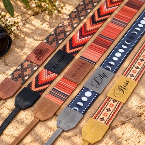 May include: Five camera straps with distinct patterns and personalized leather tabs. Patterns include brown diamonds, red geometric chevrons, orange stripes, moon phases on blue, and orange floral motifs. Tabs feature names Thomas, Harris, Matilda, Lily, and Ross.