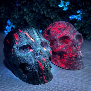 May include: Two crystal skull sculptures, one green with red streaks and one red with black streaks. The skulls are sitting on a wooden surface.