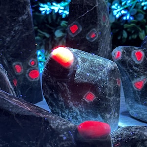 May include: Close-up of a collection of dark gray and black stones with red inclusions. The stones are polished and have a smooth surface. The red inclusions are scattered throughout the stones and appear to be glowing.