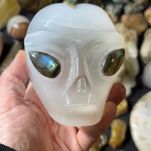 May include: White alabaster alien head sculpture with iridescent labradorite eyes. The sculpture is carved in the shape of an alien head with a smooth, white surface. The eyes are made of labradorite, a gemstone that shimmers with iridescent colors.