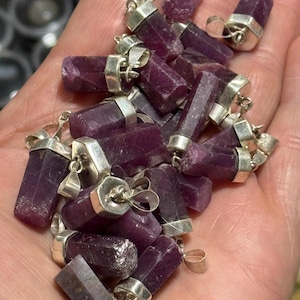 May include: A collection of purple gemstone pendants with silver bail settings. The pendants are all different shapes and sizes, but they are all roughly rectangular. The gemstones are a deep purple color and have a slightly iridescent sheen.