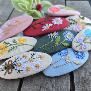 Op de afbeelding: Een verzameling geborduurde haarclips in verschillende kleuren, waaronder roze, rood, groen en blauw. Elke clip heeft een ander bloemmotief, zoals madeliefjes, rozen en andere wilde bloemen. De clips zijn ovaal en gemaakt van stof.