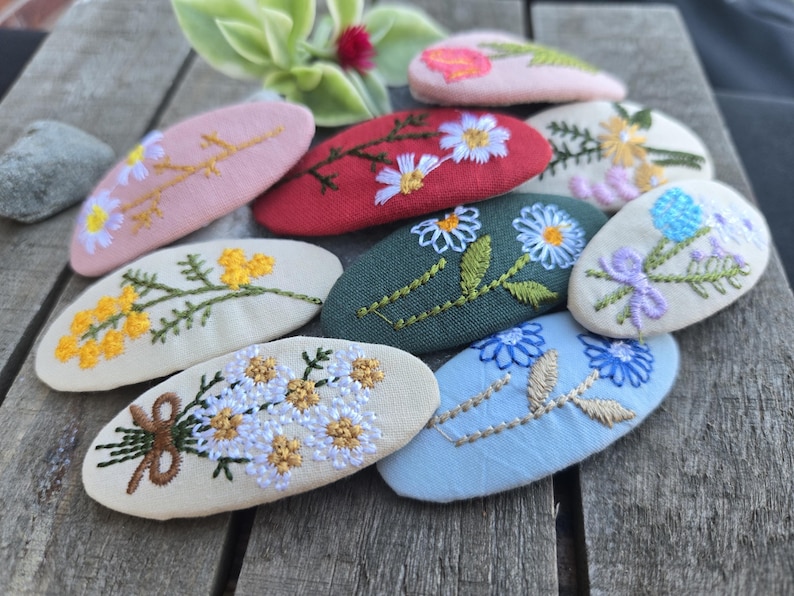 Op de afbeelding: Een verzameling geborduurde haarclips in verschillende kleuren, waaronder roze, rood, groen en blauw. Elke clip heeft een ander bloemmotief, zoals madeliefjes, rozen en andere wilde bloemen. De clips zijn ovaal en gemaakt van stof.