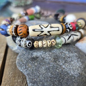 May include: Two beaded bracelets with a variety of colorful beads, including orange, blue, and white. One bracelet features a large, cream-colored bead with a black design. The bracelets rest on a gray stone.