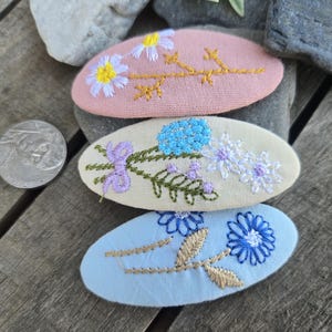 Op de afbeelding: Drie ovaalvormige, geborduurde haarclips in pasteltinten. De bovenste clip is roze met een madeliefjesontwerp, de middelste is cr&egrave;mekleurig met blauwe en paarse bloemen en de onderste is lichtblauw met een blauwe bloem en gouden accenten.