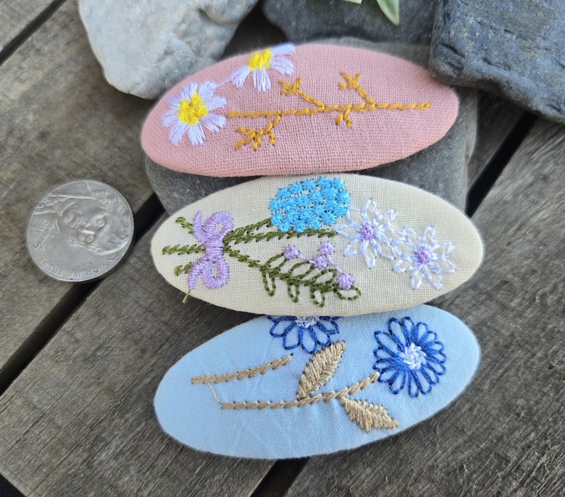 Op de afbeelding: Drie ovaalvormige, geborduurde haarclips in pasteltinten. De bovenste clip is roze met een madeliefjesontwerp, de middelste is cr&egrave;mekleurig met blauwe en paarse bloemen en de onderste is lichtblauw met een blauwe bloem en gouden accenten.