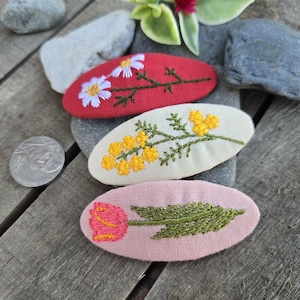 Op de afbeelding: Drie geborduurde haarclips met bloemmotieven. E&eacute;n rode clip heeft witte madeliefjes, een cr&egrave;mekleurige clip heeft gele mimosa en een roze clip toont een roze tulp. De clips zijn ovaal van vorm en liggen op een houten oppervlak.