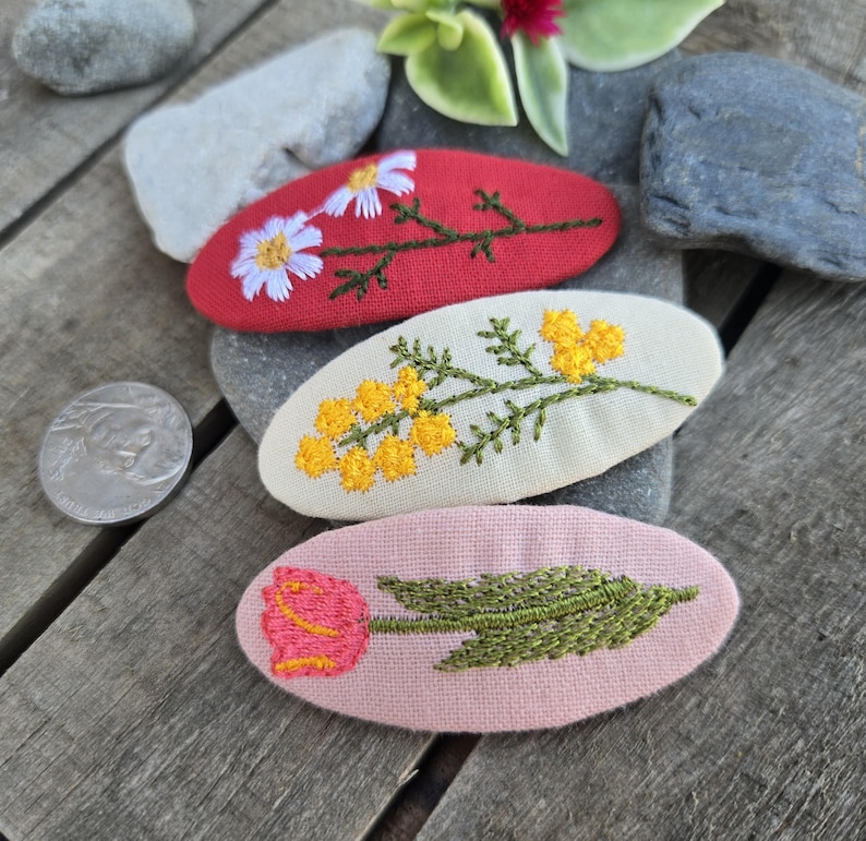 Op de afbeelding: Drie geborduurde haarclips met bloemmotieven. E&eacute;n rode clip heeft witte madeliefjes, een cr&egrave;mekleurige clip heeft gele mimosa en een roze clip toont een roze tulp. De clips zijn ovaal van vorm en liggen op een houten oppervlak.