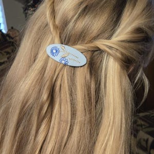 Op de afbeelding: Een lichtblauwe ovale haarclip met een bloemenontwerp. De clip heeft geborduurde blauwe bloemen en goudkleurige bladeren. De clip wordt gebruikt om blond haar vast te houden.