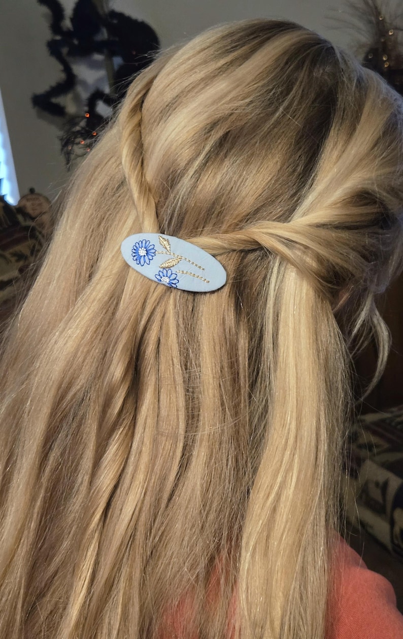 Op de afbeelding: Een lichtblauwe ovale haarclip met een bloemenontwerp. De clip heeft geborduurde blauwe bloemen en goudkleurige bladeren. De clip wordt gebruikt om blond haar vast te houden.