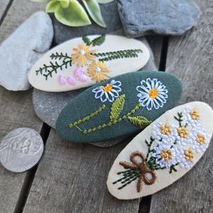 Op de afbeelding: Drie geborduurde haarclips met bloemmotieven. E&eacute;n clip is groen met witte madeliefjes, een andere is cr&egrave;mekleurig met gele en roze bloemen, en de derde is cr&egrave;mekleurig met een boeket witte en gele bloemen.