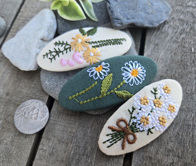 Op de afbeelding: Drie geborduurde haarclips met bloemmotieven. E&eacute;n clip is groen met witte madeliefjes, een andere is cr&egrave;mekleurig met gele en roze bloemen, en de derde is cr&egrave;mekleurig met een boeket witte en gele bloemen.