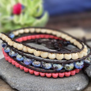 May include: A multi-strand wrap bracelet featuring dark brown leather cord woven with three distinct rows of beads. The rows consist of smooth cream-colored beads, blue and white marbled glass beads, and small solid coral-pink beads.