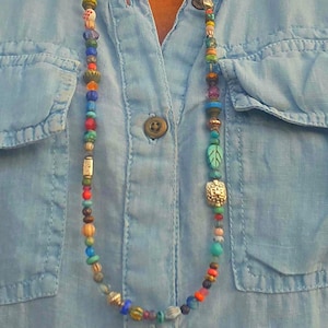 Op de afbeelding: Een lange kralenketting met een verscheidenheid aan kleurrijke kralen, waaronder rood, blauw, groen en goud. De ketting wordt gedragen over een lichtblauw overhemd met knopen. De kralen hebben verschillende vormen en maten, wat een uniek en artistiek ontwerp creëert.