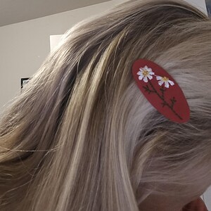 Op de afbeelding: Een rode ovale haarclip met een bloemenontwerp. De clip heeft twee witte madeliefjes met gele centra en groene stelen. De clip is geplaatst in blond haar.