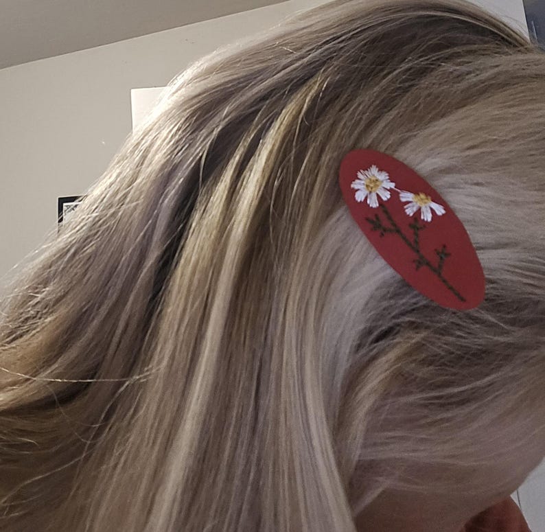 Op de afbeelding: Een rode ovale haarclip met een bloemenontwerp. De clip heeft twee witte madeliefjes met gele centra en groene stelen. De clip is geplaatst in blond haar.