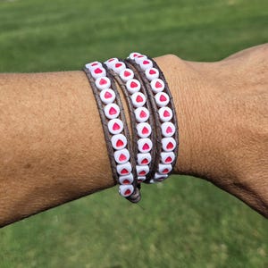 May include: Wrap bracelet with brown leather and white beads with red heart accents. The bracelet is wrapped around a wrist, showcasing the design and color contrast against the skin.