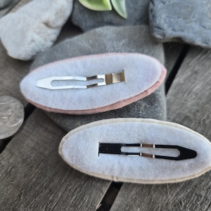 Op de afbeelding: Twee ovale haarspeldjes met metalen clips. E&eacute;n speldje heeft een roze stoffen rand, de andere een beige rand. De clips zijn zilver en zwart. Er ligt een munt naast de speldjes.
