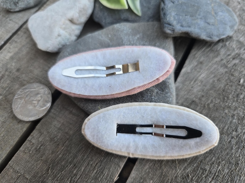 Op de afbeelding: Twee ovale haarspeldjes met metalen clips. E&eacute;n speldje heeft een roze stoffen rand, de andere een beige rand. De clips zijn zilver en zwart. Er ligt een munt naast de speldjes.