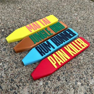 May include: Four brightly coloured bar signs, each with a cocktail name: Mai Tai, Jungle Bird, Rum Runner, and Pain Killer. The signs are yellow, brown, blue, and red, with contrasting text. Ideal for a home bar or pub setting.