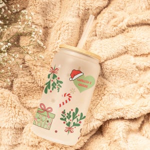 May include: A frosted glass tumbler with a bamboo lid and a straw. The tumbler features a Christmas-themed design with illustrations of holly, candy canes, presents, and a Santa hat. The text "Santacito y Chispa" is printed on the tumbler.