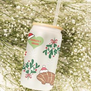 May include: A white frosted glass tumbler with a bamboo lid and a straw. The tumbler features a Christmas-themed design with snowflakes, holly, and a croissant wearing a Santa hat. The text "Cafecito y Chisne" is printed on the tumbler.