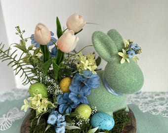Peut inclure: Une décoration de Pâques avec un lapin vert entouré de fausses fleurs, d'œufs tachetés et de verdure. L'arrangement comprend des tulipes roses claires, des fleurs bleues et jaunes, et une base recouverte de mousse, créant une ambiance printanière festive.